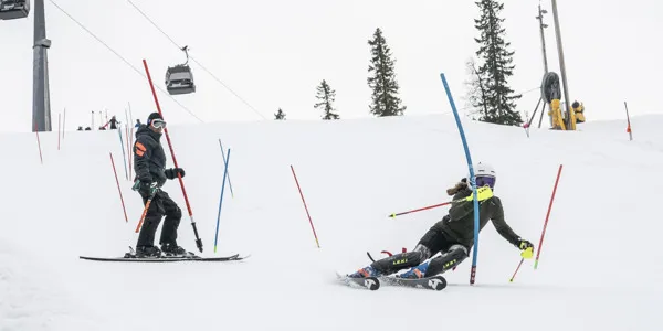A young girl training in the slalom slope, weaving between poles, with her coach standing higher up the slope.